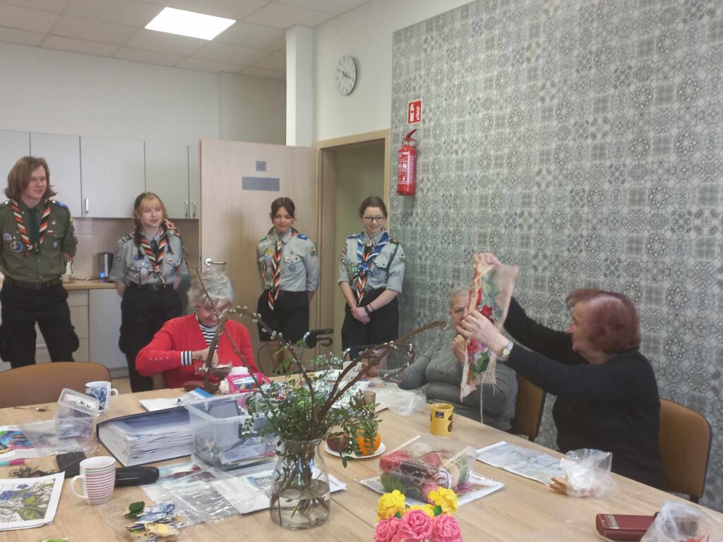 Harcerze stoją w pracowni w DDS+ i podziwiają pracę , którą pokazuje im seniorka.