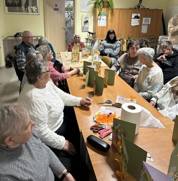 Seniorzy z DDS+ wraz z młodzieżą ze Szkoły Podstawowej w Sieniawie Żarskiej siedzą przy stole i prezentują wykonane kartki wielkanocne